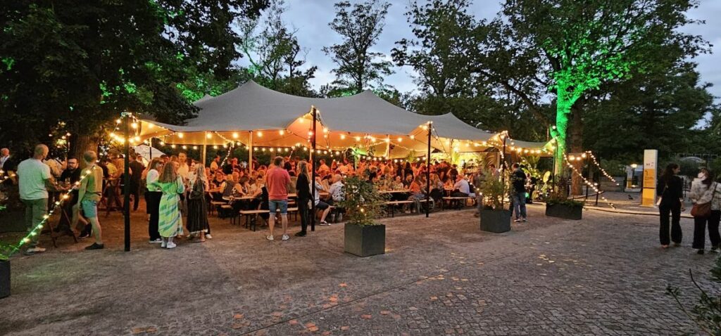 Flexzelt bei Abend mit Lichterketten, vielen Gästen an Holztischen und sommerlicher Outdoor-Veranstaltung im Grünen
